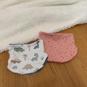 Könnte beinhalten: Zwei Baby-Lätzchen, eines mit rosa Hintergrund und kleinen weißen Herzen und das andere mit weißem Hintergrund und einem Dinosaurier-Muster in Blau, Grün und Braun.