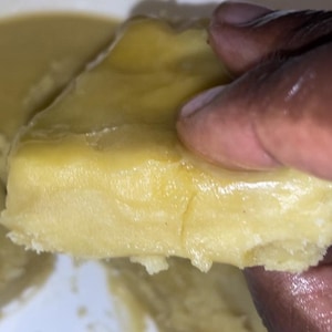 Puede incluir: Primer plano de un trozo de comida amarilla, posiblemente masa o un alimento cocinado. La comida tiene un aspecto suave y texturizado y está sostenida por una mano de piel oscura. La comida está en un cuenco blanco.