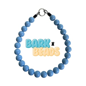 May include: A light blue beaded dog collar with a black adjustable buckle and a silver metal ring. The collar has the text "BARK x BEADS" printed on it.