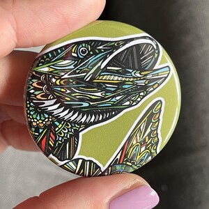 May include: A green button with a detailed black and white illustration of a fish with its mouth open. The fish is surrounded by colorful patterns.