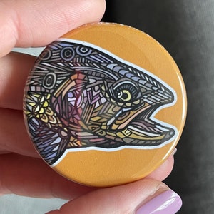 May include: A round button with a yellow background and a colorful, detailed illustration of a fish. The fish is drawn in a geometric style with intricate patterns and lines.