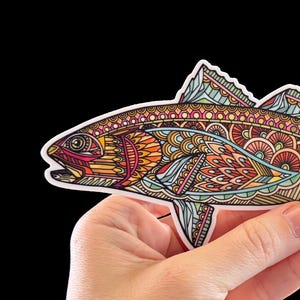 May include: A colorful sticker featuring a fish design with intricate patterns in shades of orange, red, blue, and green. The fish is held against a black background. The sticker is approximately 6 inches long.