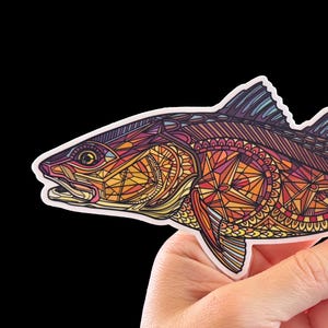 May include: A colorful sticker of a fish with intricate geometric patterns in shades of orange, yellow, purple, and blue. The fish is held in a hand against a black background. The sticker has a white border.
