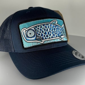 Può includere: Un cappello da camionista blu navy con un design di pesce blu e bianco ricamato sul davanti. Il cappello ha una parte posteriore in rete e un visiera curva.