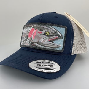 Può includere: Un cappello da camionista blu navy con una rete grigia sul retro. Il cappello presenta una toppa di una trota con una striscia rosa sul lato. Il cappello ha anche un'etichetta argentata che dice "The Authentic Yupoong Snapback Yupoong Since 1974".