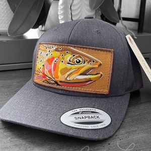 Könnte beinhalten: Eine dunkelgraue Baseballkappe mit einem braunen Patch, der eine bunte Forelle zeigt. Der Patch trägt den Text "The Authentic Yupoong Snapback Yupoong Since 1974".