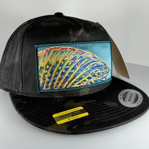 Può includere: Un cappello da camionista nero mimetico con una parte posteriore in rete nera. Il cappello presenta una toppa ricamata colorata di una pinna di pesce sul davanti. Il cappello ha anche una visiera mimetica nero e verde.