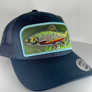 Può includere: Un cappello da camionista blu navy con schienale in rete e una toppa con una trota colorata sul davanti. La toppa è circondata da un bordo blu chiaro.
