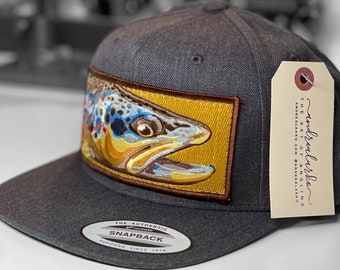 Könnte beinhalten: Eine anthrazitfarbene Snapback-Kappe mit einem gestickten Aufnäher einer Bachforelle. Der Aufnäher befindet sich auf einem goldenen Hintergrund mit braunem Rand. Die Kappe hat einen flachen Schirm und ein Etikett mit dem Text "The Art of Angling".