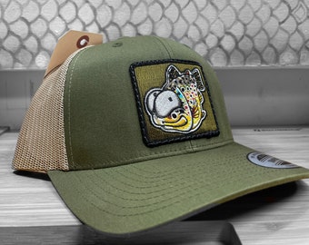 May include: A green and brown trucker hat with a patch of a cartoon trout fish. The patch has a yellow, brown, and green color scheme.