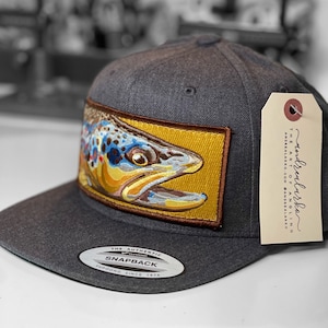 以下が含まれることがあります： チャコールグレーのスナップバックハット。茶色のマスを刺繍したパッチが付いています。パッチは茶色の縁取りのある金色の背景にあります。帽子には平らなつばがあり、「The Art of Angling」というテキストのタグが付いています。