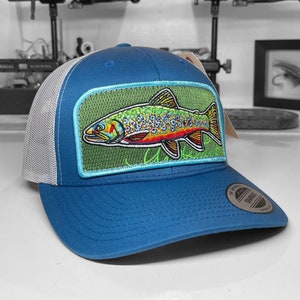 May include: A blue and white trucker hat with a green patch featuring a colorful embroidered trout. The patch has a light blue border and a green background.