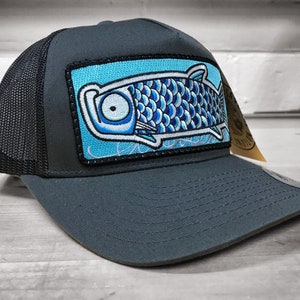 Può includere: Un cappello da camionista grigio scuro con una rete nera sul retro. Il cappello presenta una toppa ricamata blu e bianca di un pesce su uno sfondo blu.