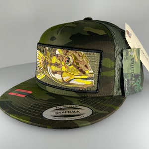 May include: A green camouflage trucker hat with a fish patch on the front. The hat has a black mesh back and a black snap closure. The hat also has a label that says "The Authentic Yupoong Snapback Since 1974".