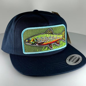 Può includere: Un cappello da baseball blu navy con una toppa ricamata di una trota marrone sul davanti. La toppa ha uno sfondo verde con cuciture blu chiaro intorno ai bordi. Il cappello ha un visore nero e un'etichetta bianca "Snapback" sul retro.