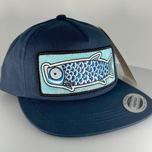 Può includere: Un cappello da baseball blu navy con una toppa di pesce blu e bianca sul davanti. La toppa è un'illustrazione stilizzata di un pesce con squame e un occhio grande. Il cappello ha una visiera curva e una chiusura a scatto.
