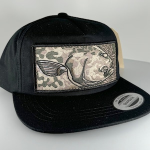 May include: Black baseball cap with a green and brown camouflage patterned fish graphic on the front. The cap has a black brim and a black adjustable strap with a snap closure. The text "The Authentic Snapback" is printed on the back of the strap.