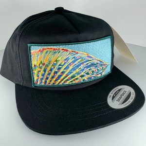 Può includere: Cappellino da baseball nero e blu con un design di pesce ricamato colorato sul davanti. Il cappello ha una cinghia regolabile nera con un'etichetta argentata che recita "The Authentic Snapback".
