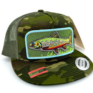 May include: A green camouflage trucker hat with a brown trout patch on the front. The patch has a blue border and features a detailed illustration of a trout with orange and yellow markings. The hat has a black mesh back and a black snap closure. The hat also has a red and white label that says "MULTICAM TROPIC" on it.
