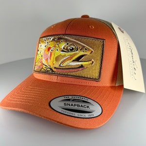 May include: Orange and brown trucker hat with a brown trout embroidered on the front. The hat has a mesh back and a snap closure. The hat also has a label that says "The Authentic Yupoong Snapback Yupoong Since 1974."