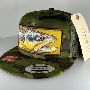 May include: A green camouflage trucker hat with a brown trout patch and a yellow background. The hat has a black mesh back and a black snap closure. The hat also has a silver metal tag that says "The Authentic Yupoong Snapback Since 1974".