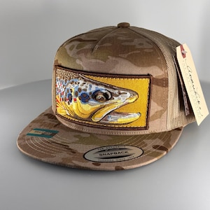 May include: A camouflage snapback hat featuring a rectangular patch with an embroidered brown trout head on a yellow background. The hat has a mesh back, a flat brim, and a sticker on the brim that reads The Authentic Snapback Yupoong Since 1974.