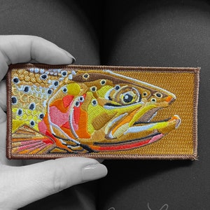 May include: A brown embroidered patch featuring a detailed illustration of a trout fish with orange, yellow, and white accents. The patch has a brown border and is perfect for adding a touch of nature to your belongings.