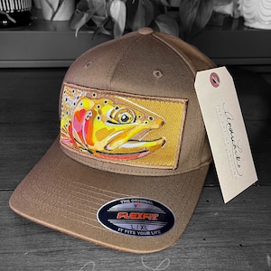 Pode incluir: Um boné de beisebol marrom com um remendo de peixe-truta bordado em marrom e amarelo na frente. O boné tem uma etiqueta Flexfit L/XL no lado. O texto "The Art of Angling" está impresso na etiqueta.