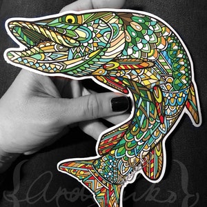 May include: A colorful, intricate, patterned sticker of a fish with a green, blue, yellow, and red design. The sticker is on a hand, and the artist's signature is visible in the lower right corner.