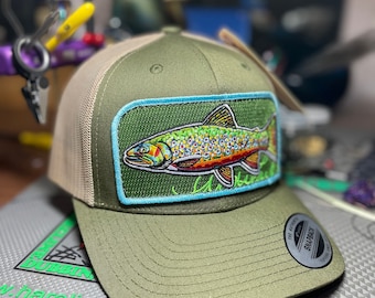 May include: A green and tan trucker hat with a brown trout embroidered patch. The patch has a blue border and a green background. The hat has a mesh back and a snap closure.