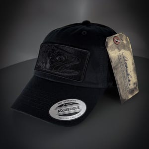 Op de afbeelding: Zwarte verstelbare baseballpet met een zwart geborduurd visontwerp aan de voorkant. De pet heeft een zilveren label met de tekst "The Authentic of Yupoong Adjustable Yupoong Since 1974".