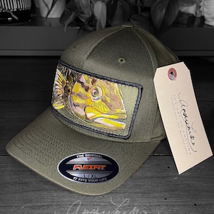 May include: A green baseball cap with a fish embroidered on the front. The cap has a Flexfit label and the text "The Art of Angling" is printed on the back.