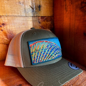 May include: A green and beige trucker hat with a colorful fish patch on the front. The patch features a blue, green, yellow, and orange design. The hat has a mesh back and a curved brim.