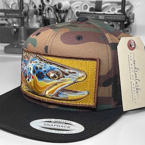 May include: A camouflage baseball cap with a black brim and a brown patch featuring a detailed illustration of a trout fish. The patch has a yellow background and brown border. The cap has a black and white label on the back that says "The Authentic Snapback Yupoong Since 1974". A brown tag with white text is attached to the side of the cap that says "andrealarka The Art of Anglin andrealarka.com @andrealarka".