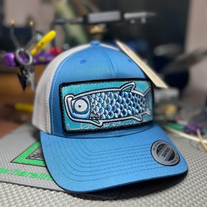 May include: A blue and white trucker hat with a blue and white fish embroidered patch on the front. The hat has a white mesh back and a black "The Authentic Snapback" label on the back.