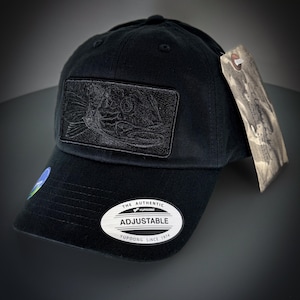 May include: Black baseball cap with a black embroidered fish design on the front. The cap has an adjustable strap with a white label that reads "The Authentic Yupoong Adjustable Yupoong Since 1974".