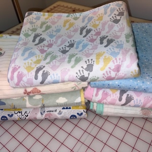 May include: A stack of folded fabric swatches featuring various patterns including colorful baby footprints, blue polka dots, yellow ducks, and nautical motifs. The textiles are layered on top of each other, displaying a variety of pastel colors and prints.