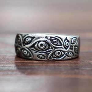 Könnte beinhalten: Ein silberner Ring mit einem sich wiederholenden Augenmuster. Der breite Ring hat ein detailliertes Muster aus zahlreichen Augen um das Band. Er liegt auf einem verschwommenen Holzgrund.