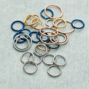 Puede incluir: Un surtido de pequeñas joyas circulares en varios colores, incluyendo plata, oro, oro rosa y azul. Algunos anillos tienen un diseño de cuerda trenzada. Las joyas están dispersas sobre una superficie gris claro.