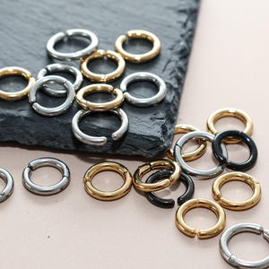 Puede incluir: Un surtido de pendientes de aro en oro, plata y negro. Los pendientes están dispuestos sobre una superficie texturizada de color gris oscuro y un fondo rosa claro. Los pendientes son de varios tamaños y estilos, con un cierre de bisagra.