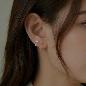 May include: Close-up of a person's ear adorned with delicate gold earrings. The ear features two small, sparkling stud earrings and a gold hoop earring. The earrings are set against the backdrop of the person's skin and dark hair.