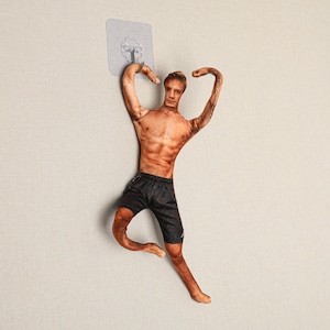 May include: A novelty wall hook shaped like a muscular man in black shorts. The hook is attached to a clear adhesive pad for easy mounting. The man's arms are raised, and his body is tanned.