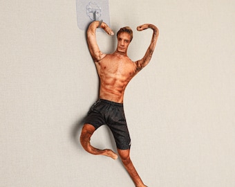 May include: A novelty wall hook shaped like a muscular man in black shorts. The hook is attached to a clear adhesive pad for easy mounting. The man's arms are raised, and his body is tanned.