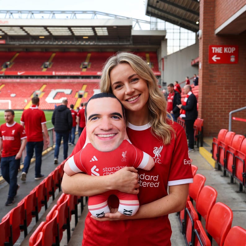 May include: A red plush doll in the shape of a soccer player, wearing a red jersey with white accents. The doll has a smiling face and is being held in front of a stadium.
