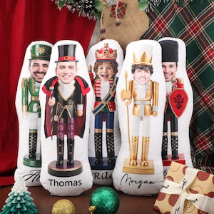 May include: Five custom-shaped plush pillows featuring printed nutcracker soldier uniforms with human faces superimposed on the heads. Each pillow displays a unique nutcracker outfit and a name printed in black script at the bottom base.
