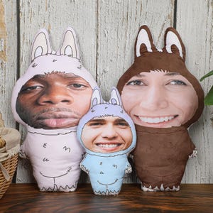 Puede incluir: Tres cojines de peluche personalizados con forma de personajes de dibujos animados, cada uno con la cara de una persona. Los cojines son de colores pastel: rosa, azul y marrón. Los cojines tienen orejas de conejo y están sobre una superficie de madera.