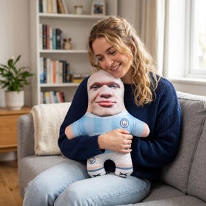 May include: A plush doll featuring a realistic face print, wearing a light blue and white soccer jersey. The doll has a round head and a smiling expression. The doll is being held by a person.