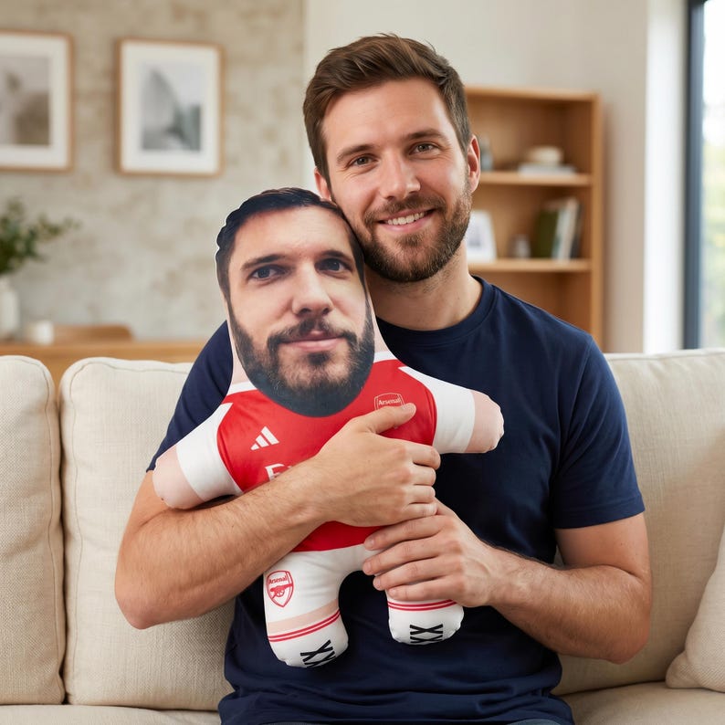 May include: A plush doll featuring a printed face and a red and white soccer jersey. The jersey has the Arsenal logo and Adidas branding. The doll is held by a person wearing a navy blue shirt.