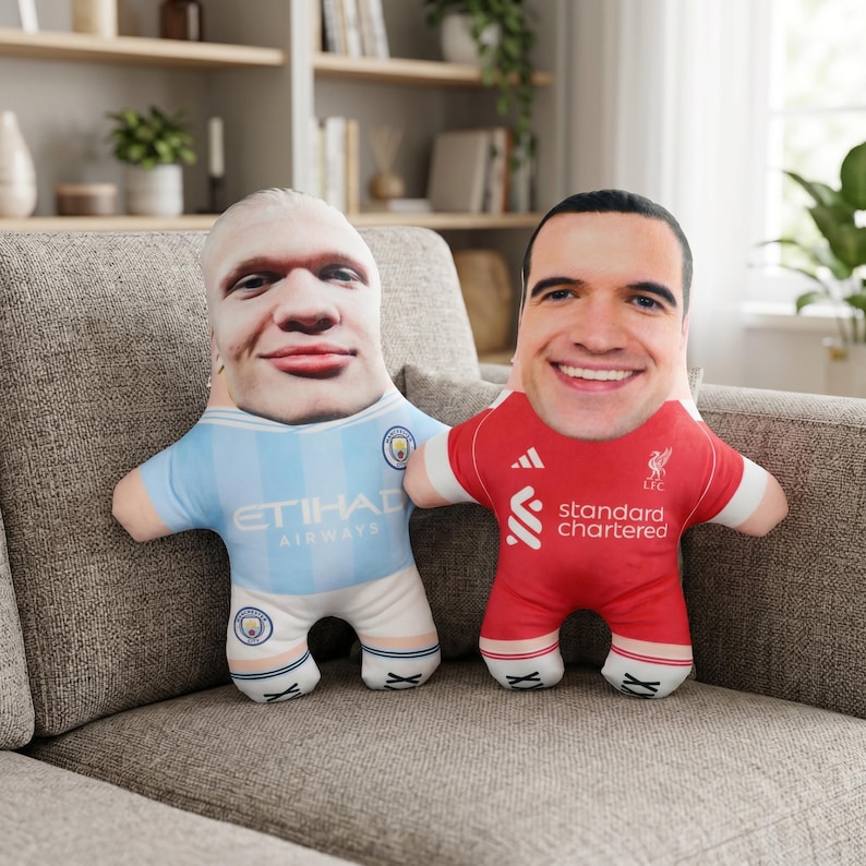 May include: Two plush dolls shaped like figures, each with a person's face printed on the head. One doll wears a light blue jersey with "Etihad Airways" printed on it. The other doll wears a red jersey with "Standard Chartered" printed on it.