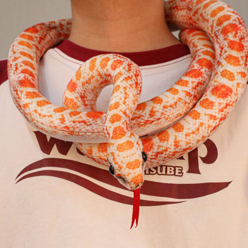 May include: A plush snake toy in shades of orange and white, coiled around a person's neck. The snake has a red tongue and black eyes. The person is wearing a white t-shirt with a maroon collar and the word "SUBE" printed on it.
